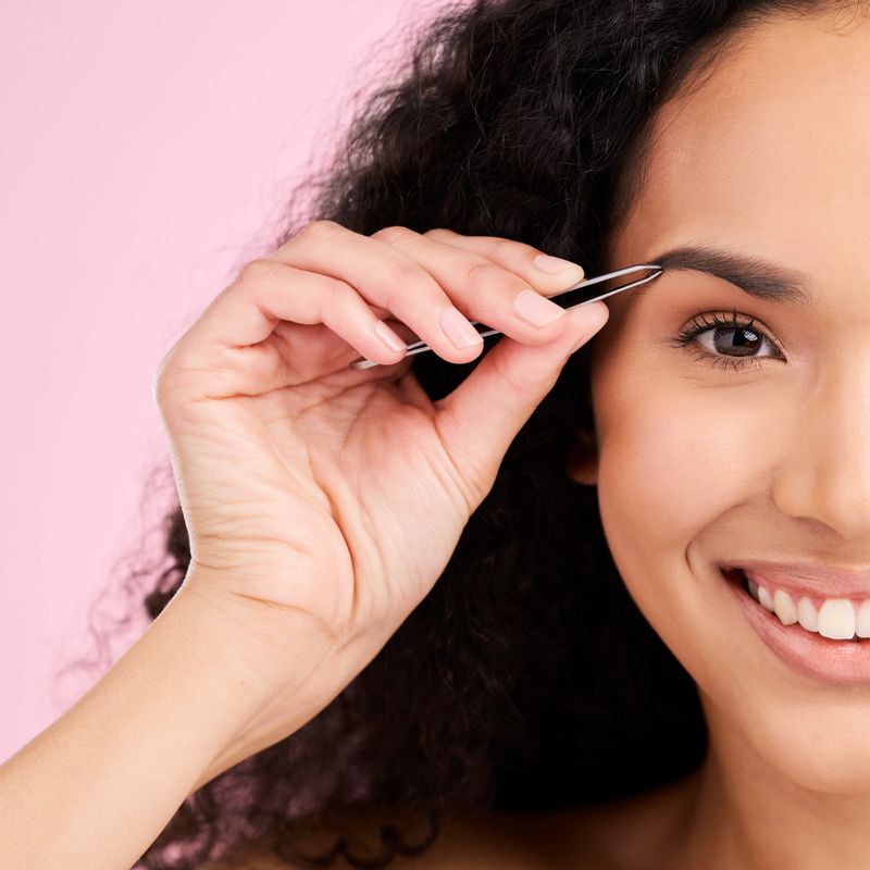 Face, happy and woman with tweezer for eyebrow, beauty and grooming isolated on a pink background in studio. Natural portrait, facial plucking and hair removal of model for wellness and aesthetic