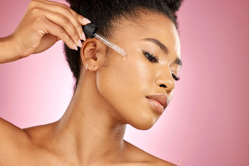 Black woman, oil and serum on face, skincare shine or aesthetic dermatology on pink background in studio. Female model, facial cosmetics and dropper of hyaluronic acid, liquid collagen or beauty glow