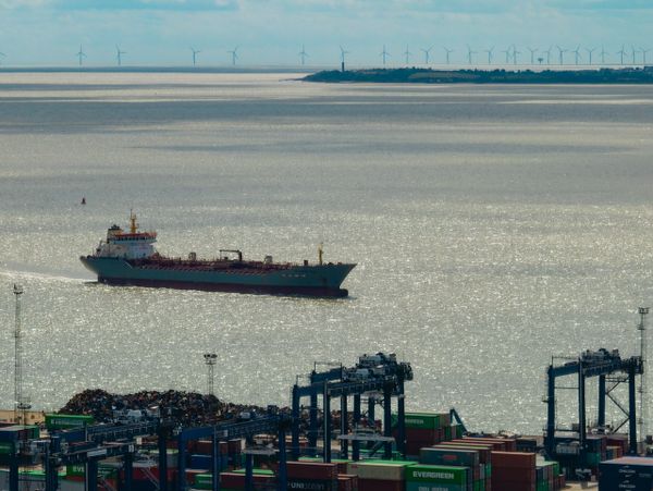 Cargo ship sailing near a busy container port with wind turbines in the background.