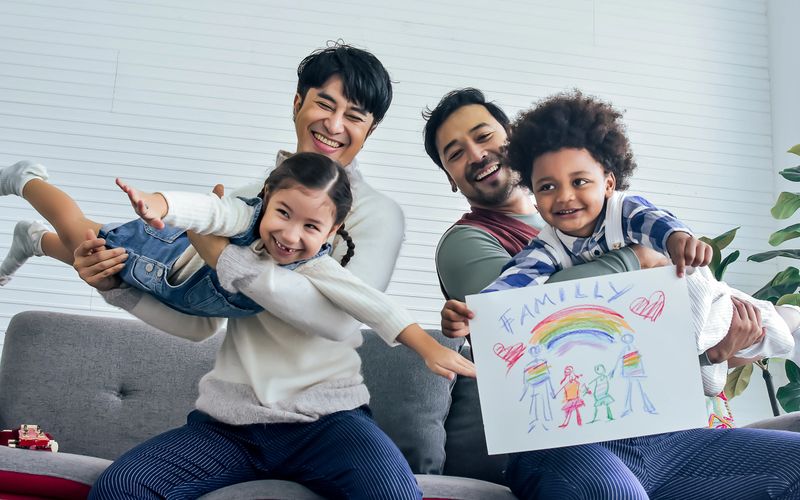 Male gay taking care adopted children who are happy diverse little Caucasian girl and African boy, playing with fun, drawing family picture, sitting on sofa in living room at home. LGBT, kids Concept