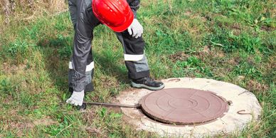 Worker in safety gear opening a manhole cover in a grassy area.
