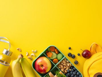 Healthy lunchbox with fruits, nuts, and vegetables on a yellow background.