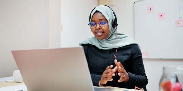 Woman in hijab wearing headset, speaking on a video call at her laptop.