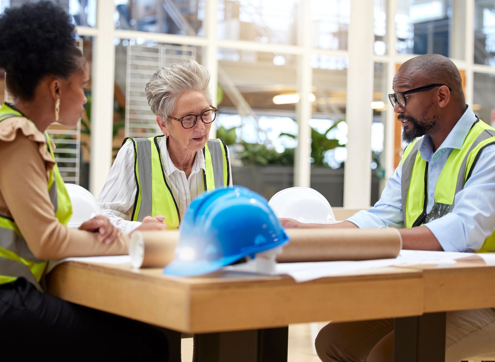 Three engineers in safety vests discuss plans at a table with helmets.