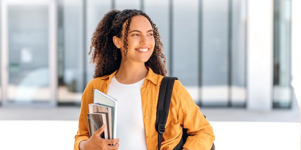 Smiling student with curly hair holding notebooks and wearing a yellow shirt and backpack.