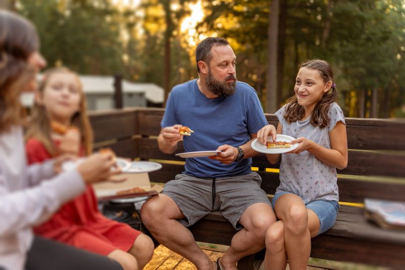 A happy family with two daughters has fun eating takeout pizza for dinner together outside on the back patio of their home on a warm and sunny evening.