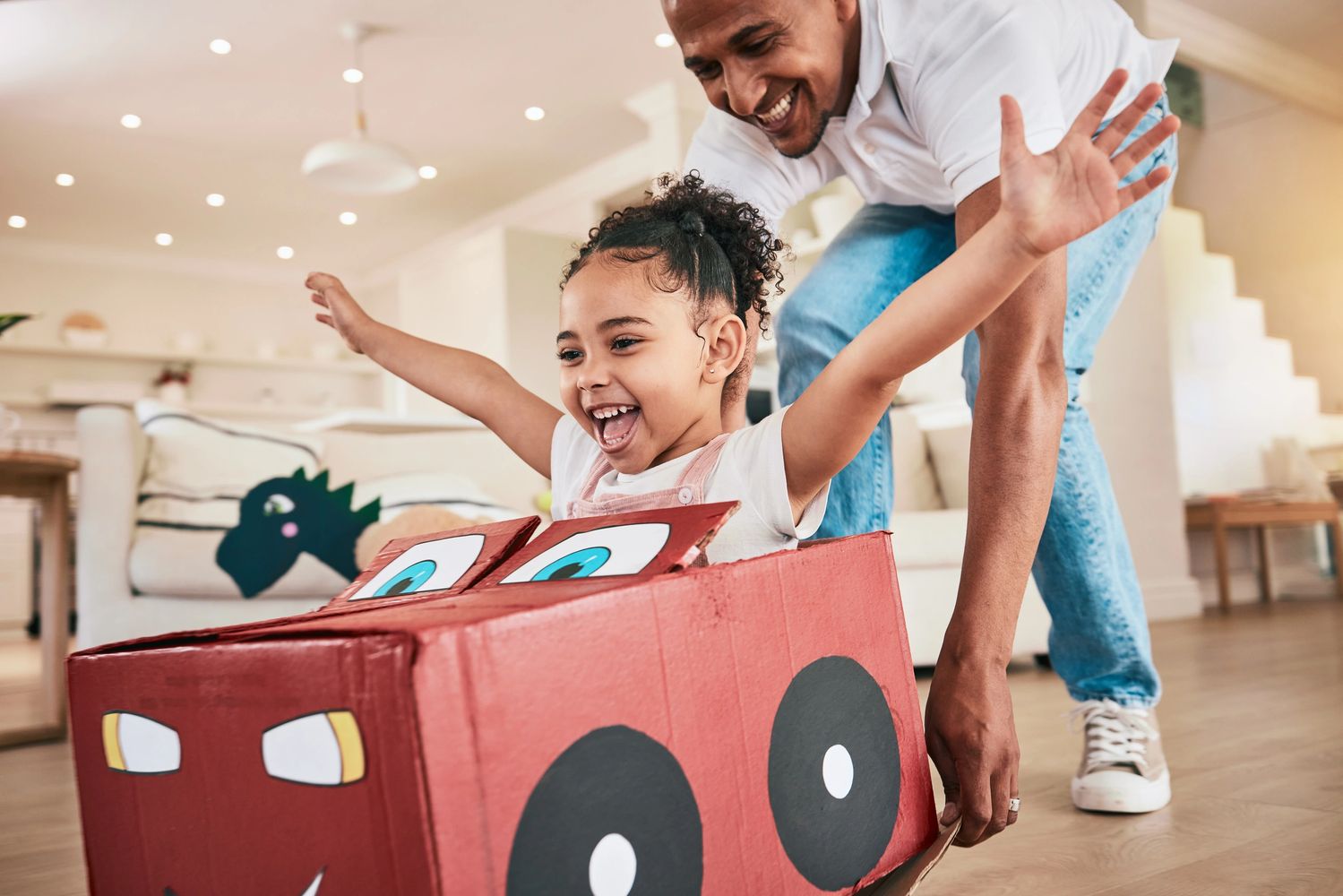 A joyful father pushes his daughter in a homemade cardboard car indoors.