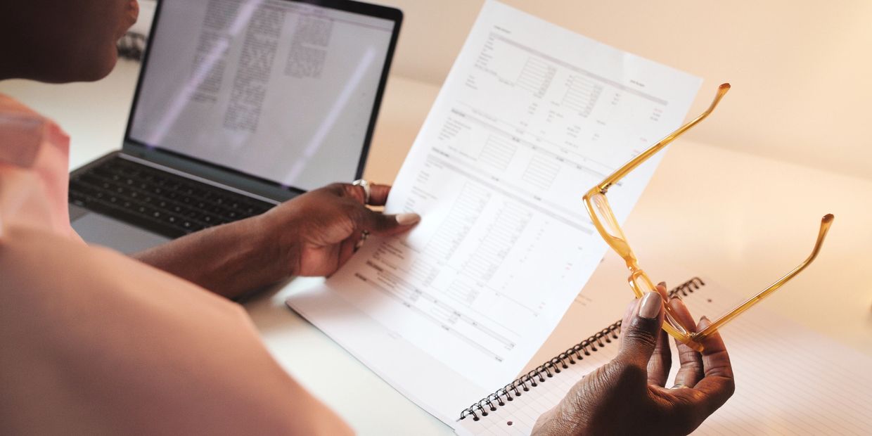 Person reviewing a document while holding glasses near a laptop and notebook.