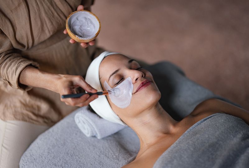 Close up shot of a beautiful smiling woman lying with her eyes closed while another woman is applying a face mask on her in the spa center. She is relaxed and enjoys the treatment.