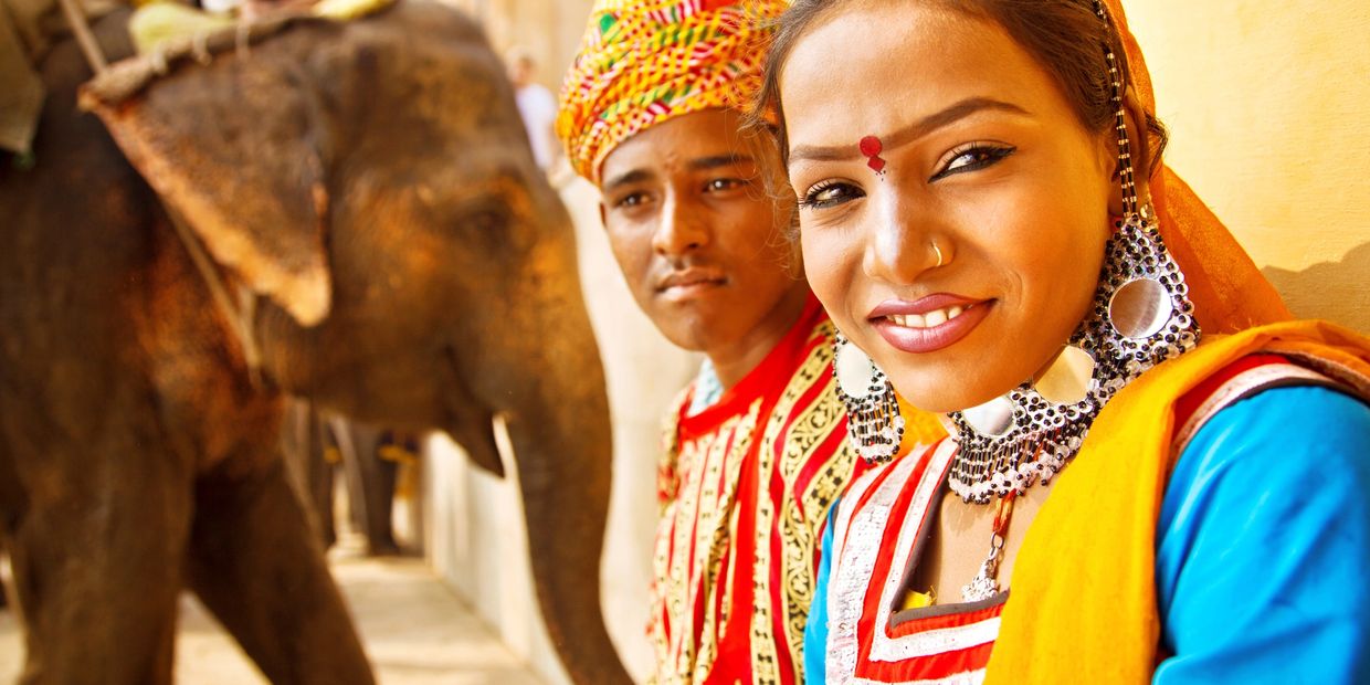 A smiling woman and man in traditional attire with an elephant in the background.