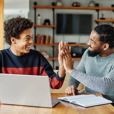 Two men happily high-five while working on a laptop and notebook at a table.