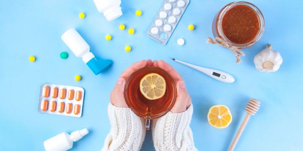 Hands holding tea with lemon surrounded by medicine and natural remedies.
