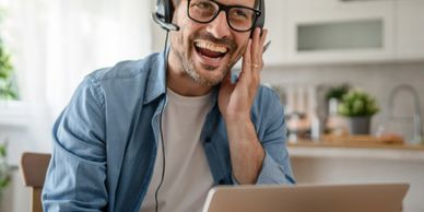 Man wearing headset, laughing while working on laptop at home.