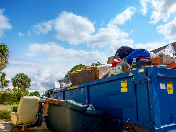A large blue dumpster overflowing with furniture and household items under a partly cloudy sky.