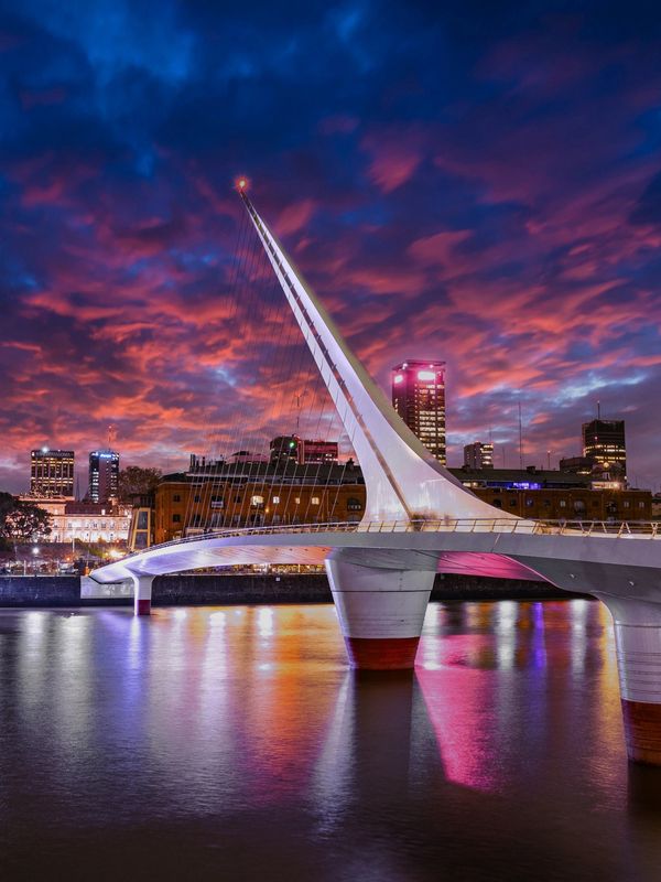 Modern white bridge lit against a vibrant sunset sky over a city river.