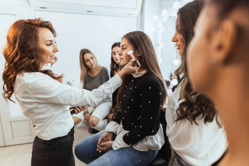 Girls are attending the make up masterclass. The educator is showing them on the model how to do glam makeup.