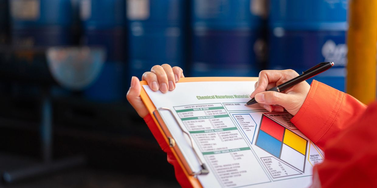 Worker filling out a chemical hazardous material checklist in front of blue barrels.