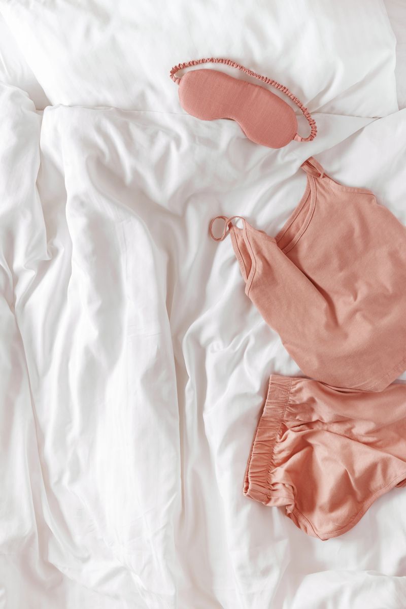 Pink woman pajamas and sleep eye mask on bed. Top view summer pyjama for sleeping. Aesthetic lifestyle flat lay photo, singlet and shorts peach colored, comfort ans stylish wear or clothes, copy space
