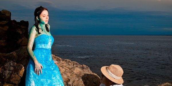 Woman in blue dress stands on rocks by the sea at dusk.