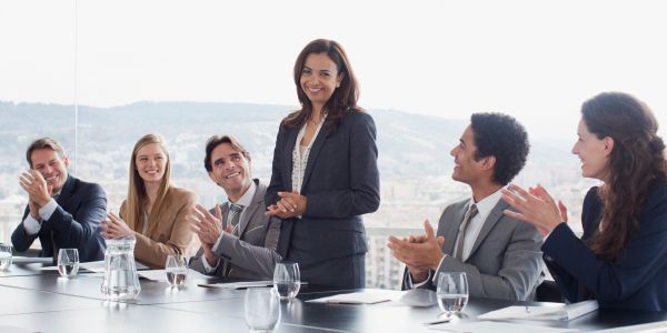 Businesswoman standing and speaking while colleagues applaud.