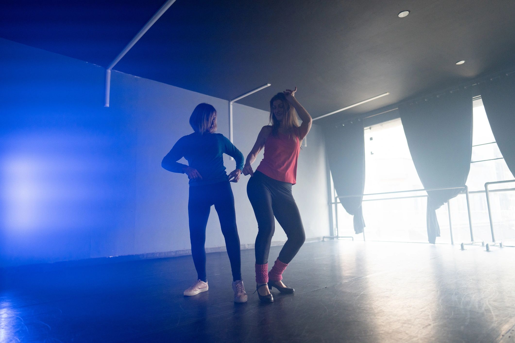Two women dancing in a dimly lit room with blue and natural light.