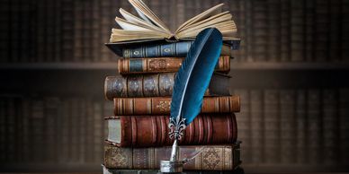 An open book atop of several closed books. A blue quill sits in an ink jar in front of the books. 