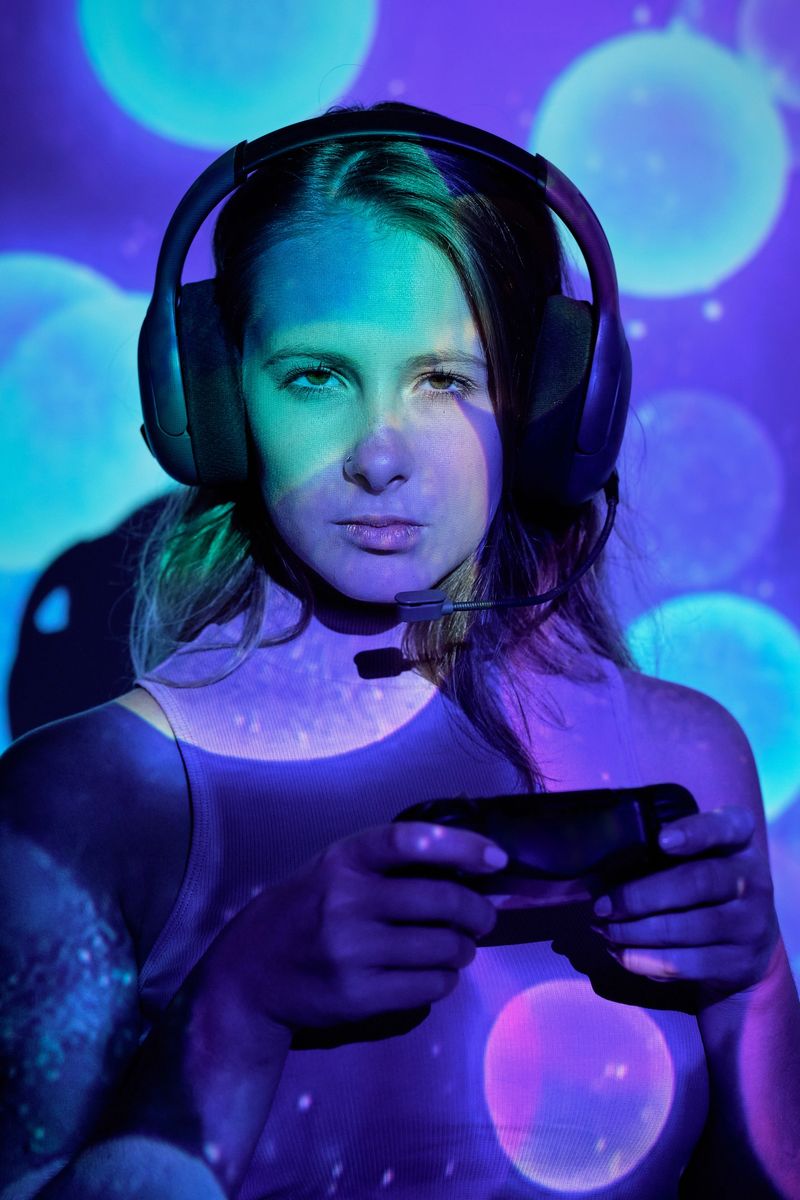 Young woman with headphones and controller looking at camera with serious face while playing difficult videogame under colorful illumination