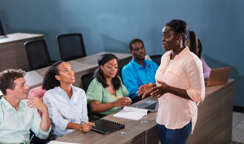 A mature African-American woman teaching a seminar or training class to a multiracial group of adults. The students are sitting in a row at a long table, with their note pads and laptop computers.