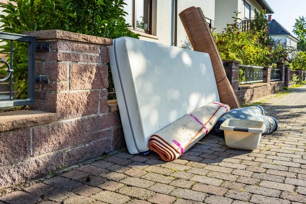 Discarded mattress, rugs, and a plastic tub on a sidewalk outside a house.