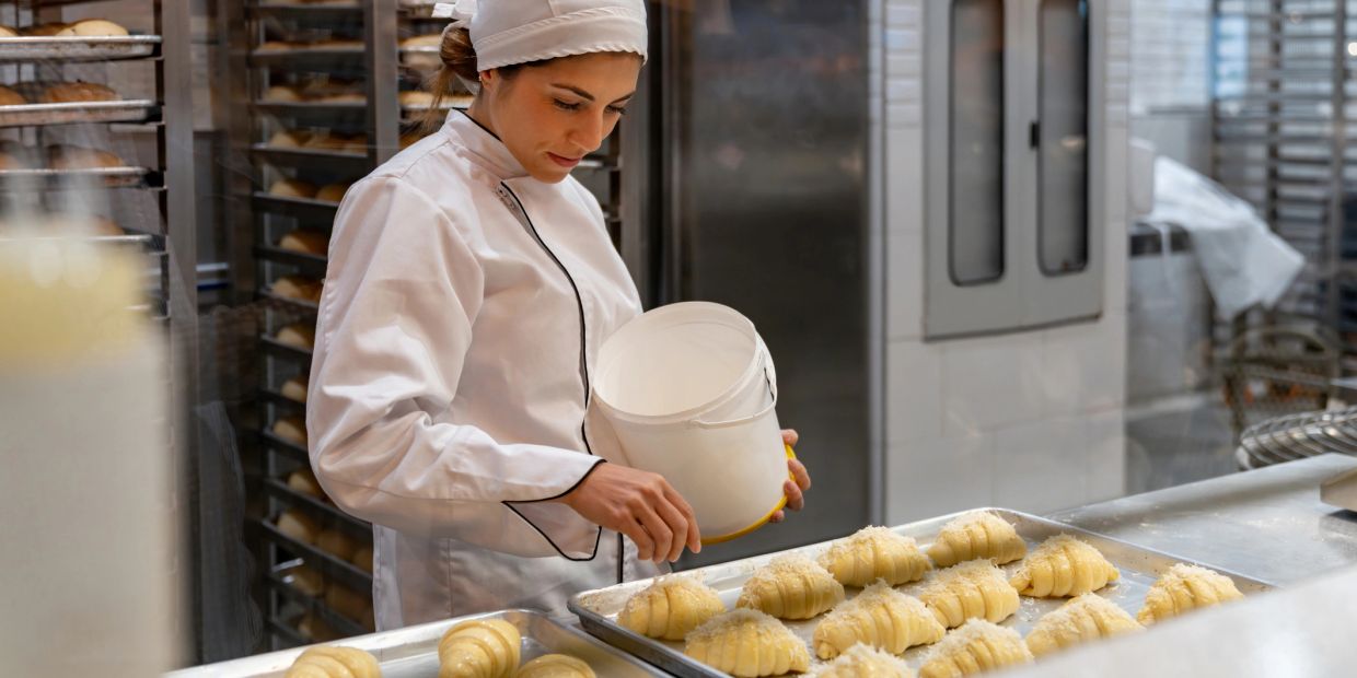 Baker sprinkling ingredients on croissants in a commercial kitchen.