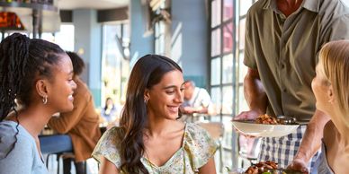 Three women happily receiving food from a waiter in a bright restaurant.