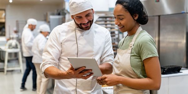 Two bakers discussing a tablet in a busy bakery kitchen.