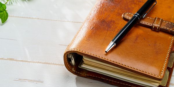 Brown leather journal with a pen on a white wooden table.
