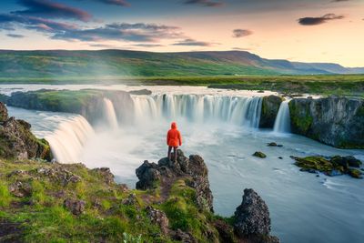 Person in red jacket stands on rock overlooking a wide waterfall at sunset.