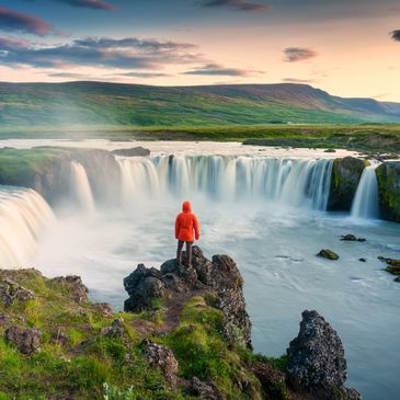A man in a red coat stands overlooking powerful Icelandic waterfalls under a cloudless sky.