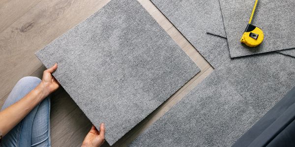 Person installing gray carpet tiles on wooden floor with measuring tape nearby.