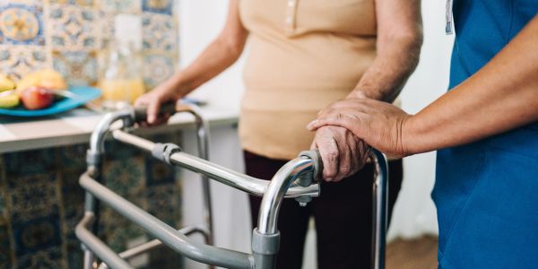 A caregiver supports an elderly person using a walker.