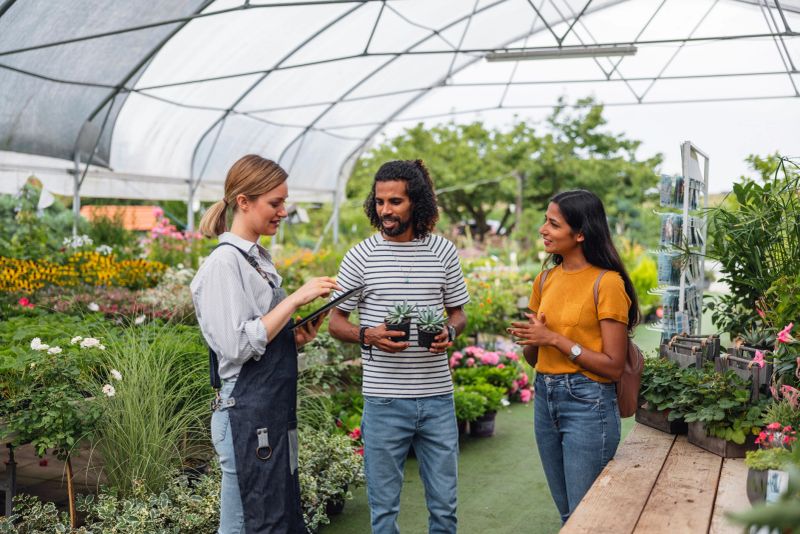 A nice and friendly adult female garden center employee having a conversation with a diverse couple wanting to buy some houseplants at a garden center. She is educating them on the care of individual plants. They are carefully listening and asking her questions. Personalized customer service.
