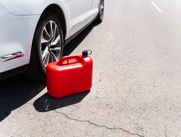 Red fuel container next to a white car's rear wheel on a sunny road.