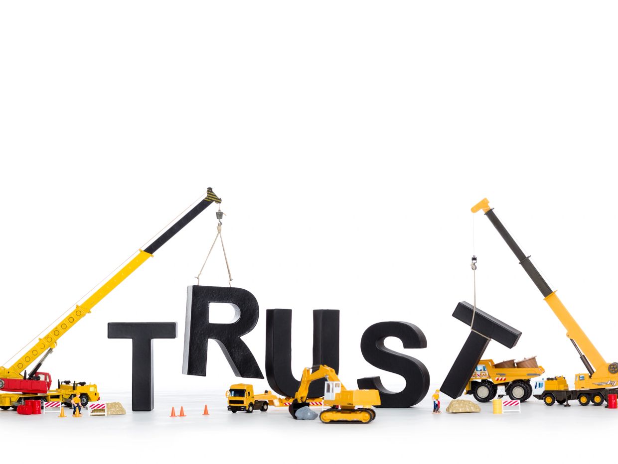 Construction vehicles assembling the word TRUST with large black letters.