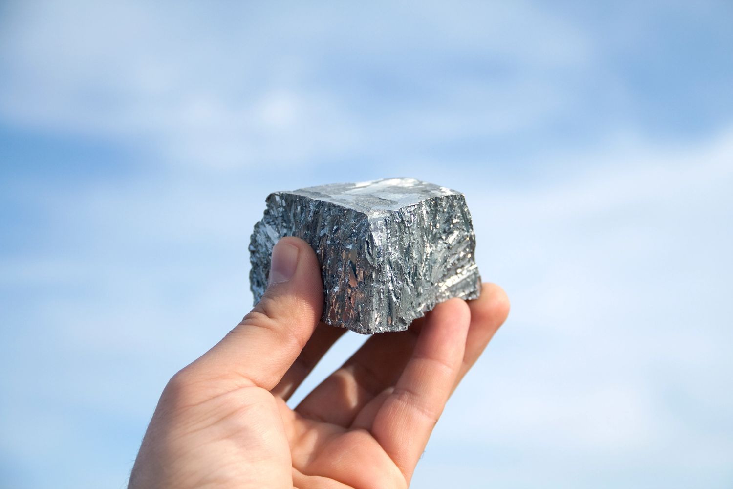 Hand holding a metallic silver rock against a clear blue sky.