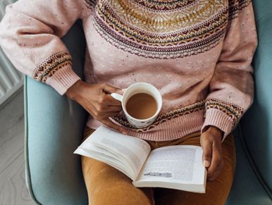 Person relaxing with a warm drink and book in a cozy sweater.