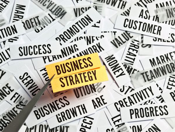 Tweezers holding a yellow note labeled 'Business Strategy' amidst scattered business-related words.