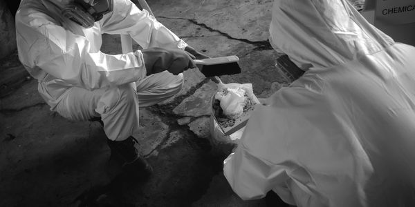 Two people in protective suits cleaning hazardous material with a brush and dustpan.