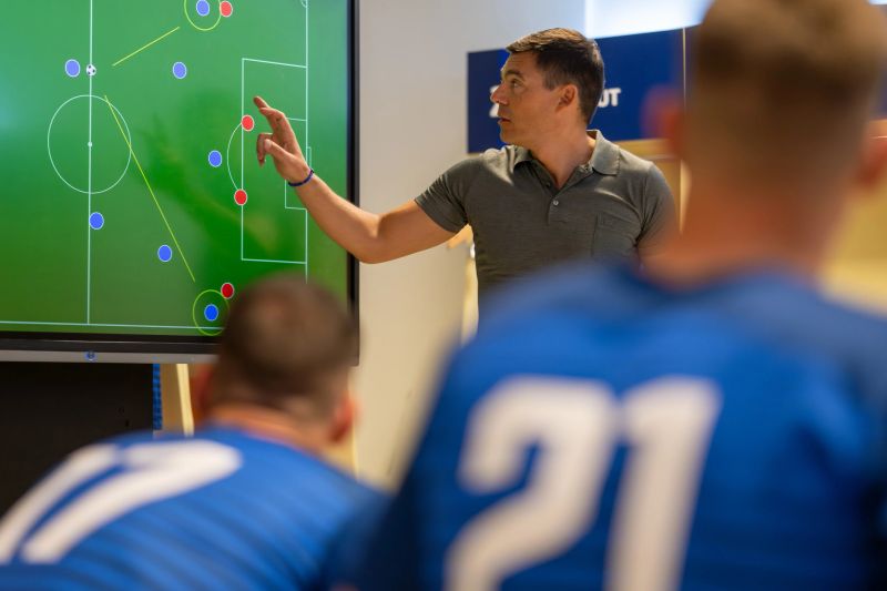 Football coach pointing while explaining game strategy on device screen to football players in dressing room.
