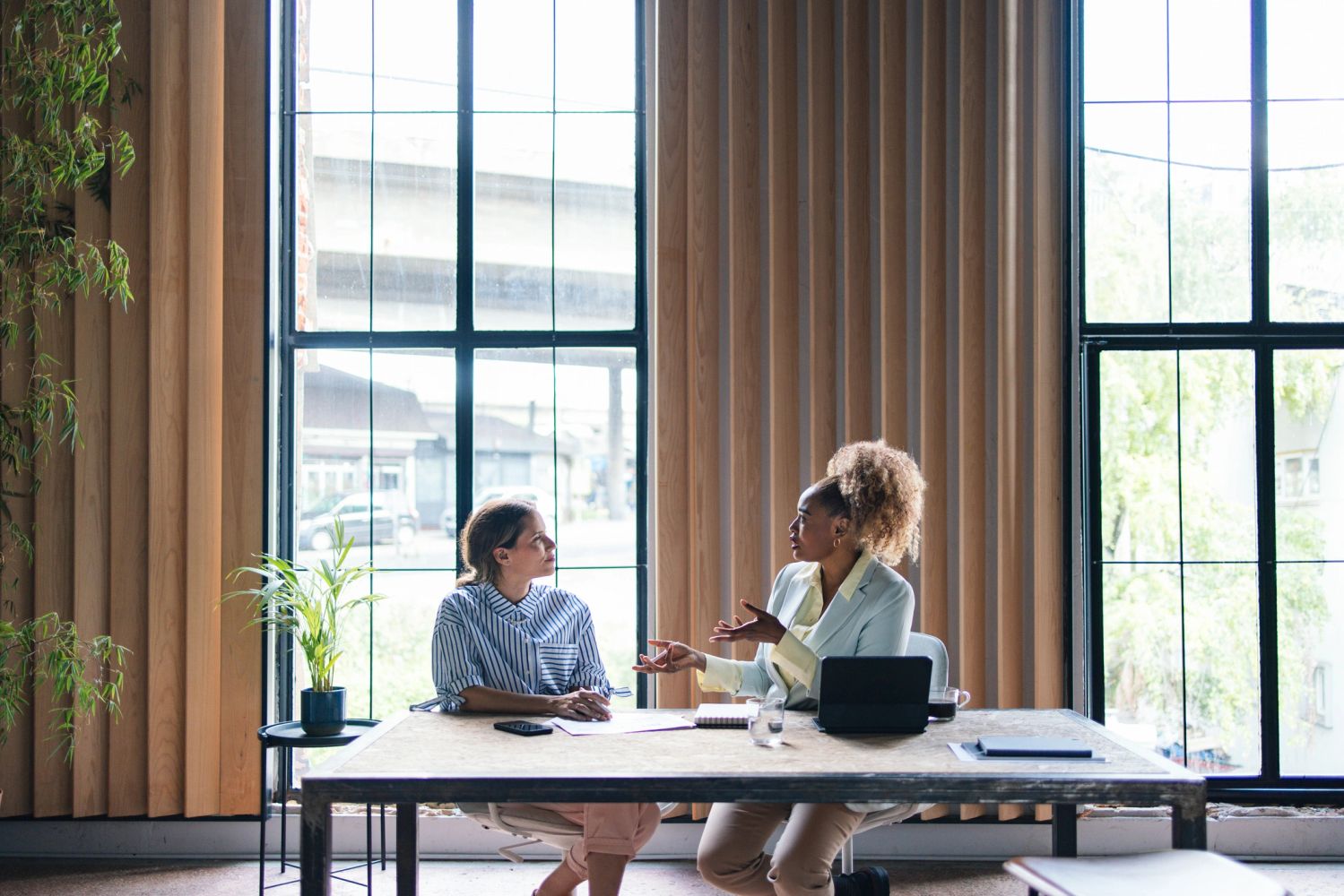 Two women having a discussion in a modern office with large windows.