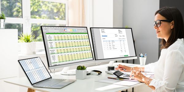 A woman working with spreadsheets and invoices on multiple screens at a bright office desk.