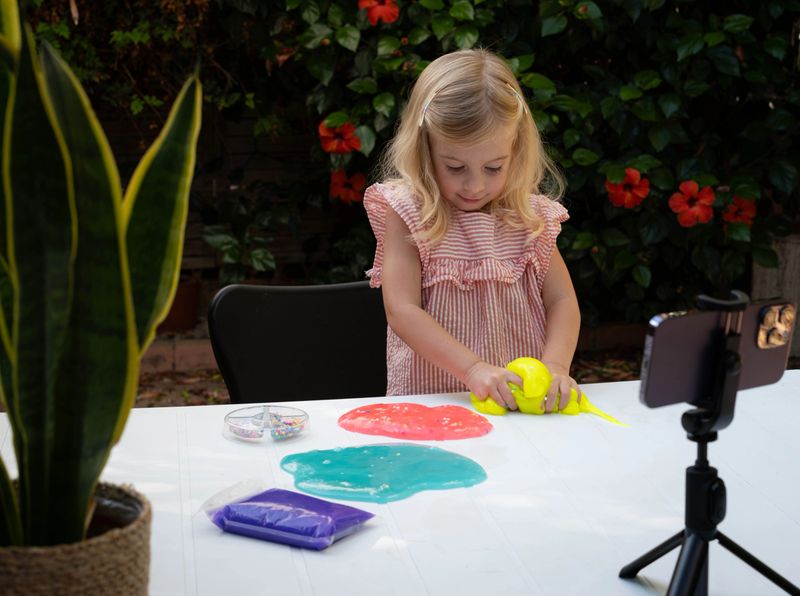 Cute blonde caucasian girl with curly hair playing with slime and watching video lesson on smart phone. Online instruction of preparing concept. Kid sensory development, creative hobby, game.
