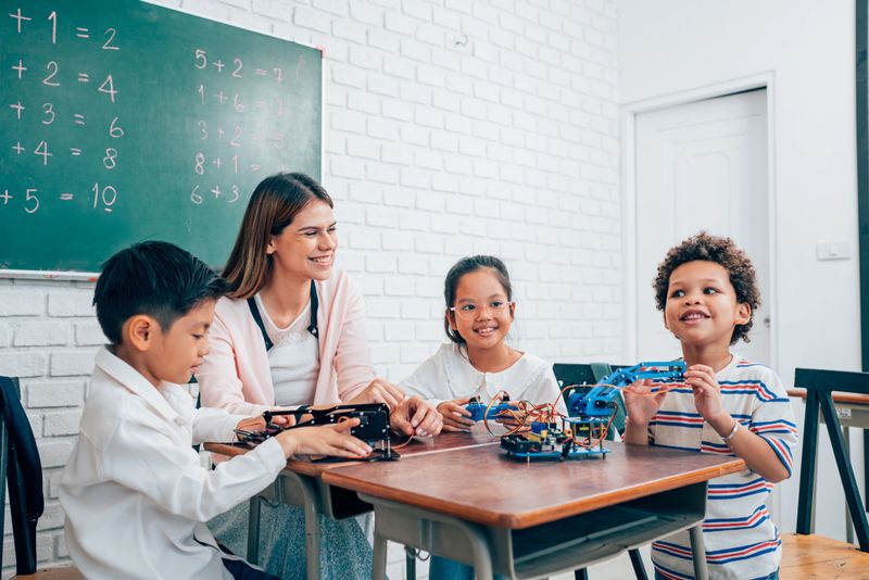 Happy school teacher and students having interesting engaging activities in class. Little children sitting around classroom table, talking and playing fun games together