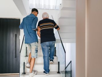 Young man helping elderly man walk upstairs inside a modern home.